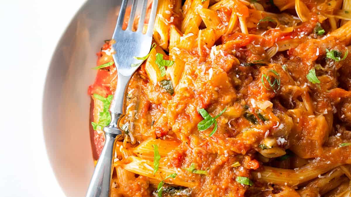 A plate of penne pasta topped with a rich tomato sauce, garnished with chopped green herbs. A fork rests on the side of the dish, offering a glimpse into delightful pasta recipes. The pasta appears to be mixed with pieces of vegetables and possibly some grated cheese.