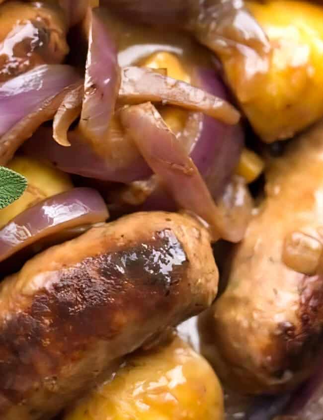 Close-up of a delicious sausage and onion dish. The sausages are surrounded by caramelized onions and garnished with fresh sage leaves. The rich, savory sauce glistens on the sausages and onions, making the dish look warm and appetizing—perfect for a slow cooker apple sausage twist.