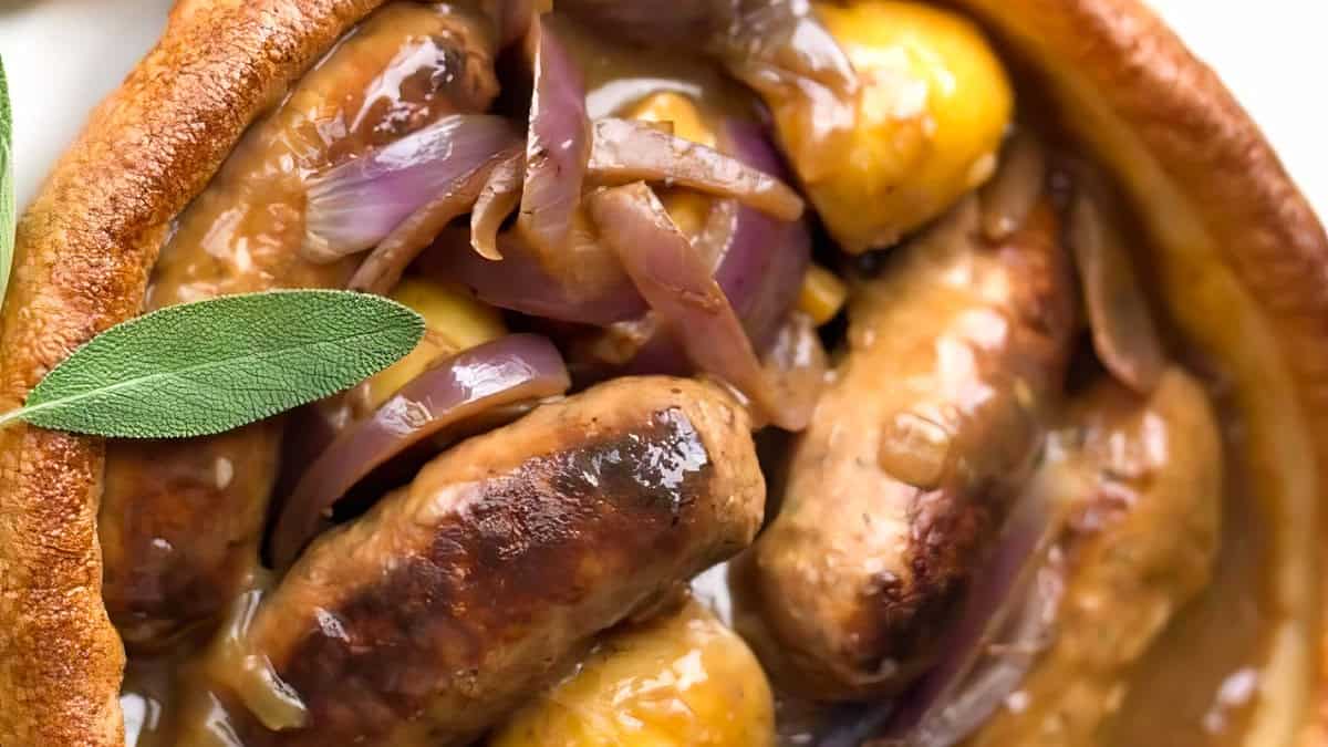 Close-up of a delicious sausage and onion dish. The sausages are surrounded by caramelized onions and garnished with fresh sage leaves. The rich, savory sauce glistens on the sausages and onions, making the dish look warm and appetizing—perfect for a slow cooker apple sausage twist.