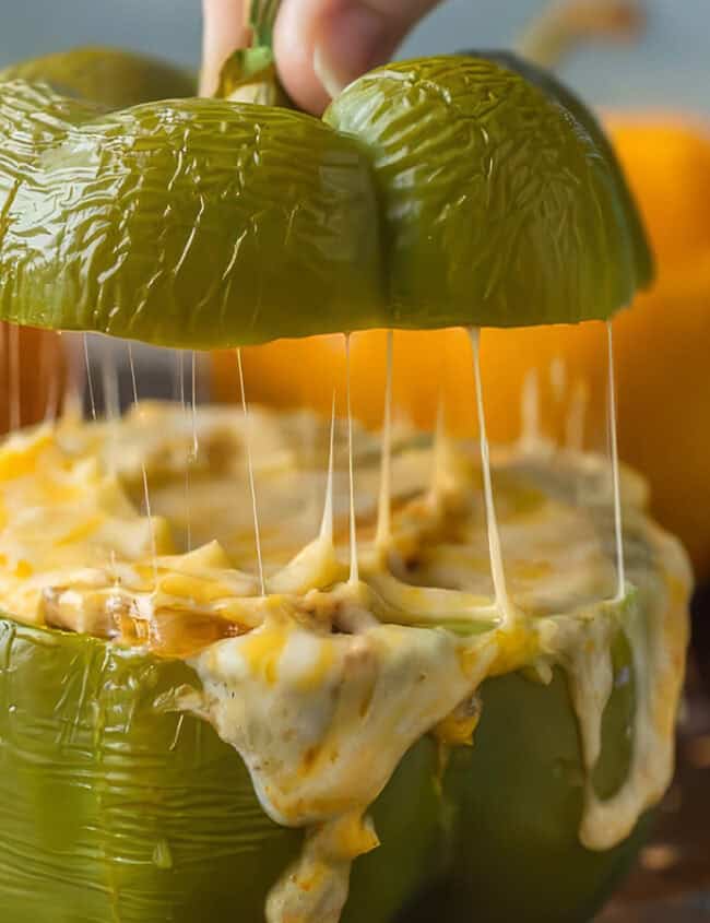 A close-up of a green bell pepper stuffed with melted cheese and other ingredients. A hand lifts the top part of the green bell pepper, revealing gooey cheese stretching. In the background, there is a yellow and a red bell pepper, also stuffed—a perfect addition to your favorite peppers recipes.