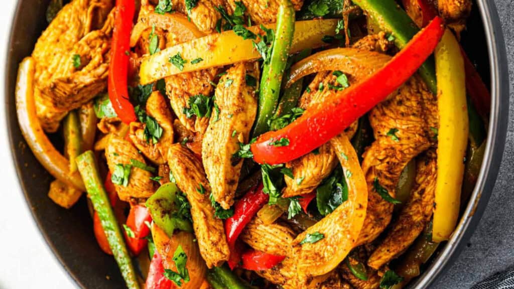 A colorful plate of chicken fajitas with thinly sliced pieces of spiced chicken, red, yellow, and green bell peppers, and onions. The dish is garnished with fresh chopped cilantro and served in a dark bowl—a perfect highlight for any peppers recipe.