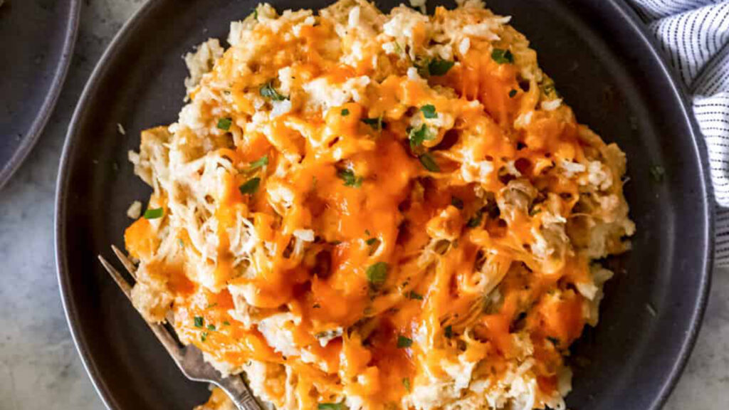 A plate of creamy chicken and rice casserole, straight from cherished rice dish recipes, is topped with a generous layer of melted cheddar cheese and garnished with chopped parsley. A fork is placed on the side of the dish. The casserole is served on a dark plate.