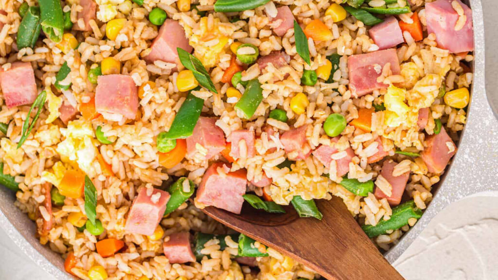 A close-up of a pan filled with fried rice. The dish contains diced ham, peas, corn, green beans, carrots, and scrambled eggs, all mixed with rice. A wooden spoon is partially submerged in the rice, suggesting it is being stirred or served—perfect for those seeking delicious rice dish recipes.