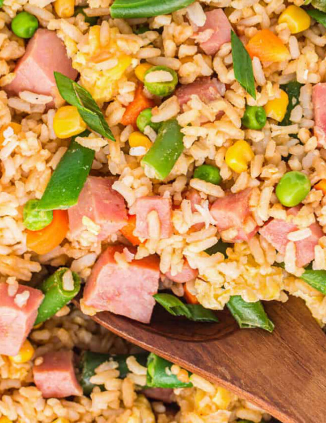 A close-up of a pan filled with fried rice. The dish contains diced ham, peas, corn, green beans, carrots, and scrambled eggs, all mixed with rice. A wooden spoon is partially submerged in the rice, suggesting it is being stirred or served—perfect for those seeking delicious rice dish recipes.