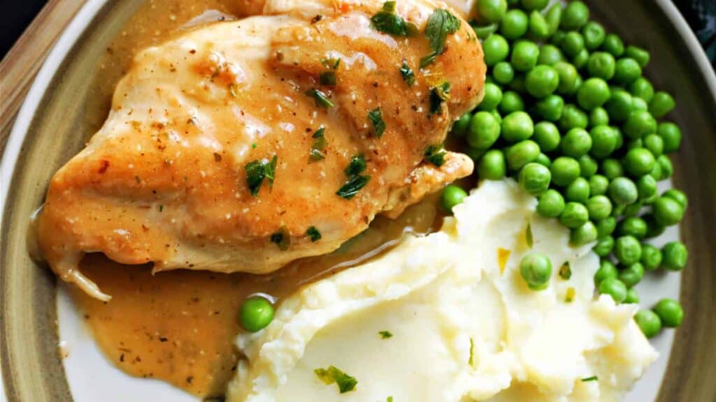 A chicken breast smothered in gravy with mash and peas