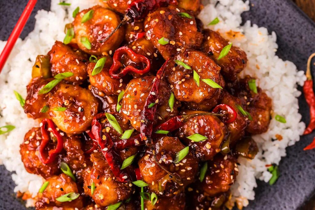 A close-up of a plate of spicy shrimp stir-fry, an Asian recipe staple, served over white rice. The shrimp is coated in a thick, glossy sauce and garnished with sliced green onions, red chili peppers, and sesame seeds. The dish is presented on a dark plate with red chopsticks partially visible.