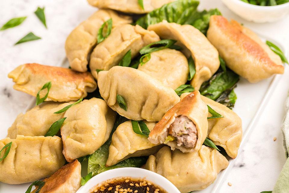 A plate of homemade dumplings filled with a meat mixture, garnished with sliced green onions. Some dumplings are placed on a bed of lettuce, and a small bowl of dipping sauce with sesame seeds is visible in the foreground, perfect for those who love exploring Asian recipes.