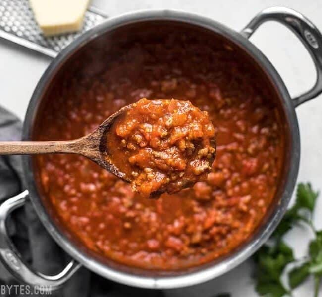 A wooden spoon holds a scoop of thick, chunky tomato sauce with ground beef above a pot filled with the delicious mixture. The pot is on a light surface with a dark fabric napkin nearby. Fresh parsley and a grater with a block of cheese are partially visible in the background, perfect for your favorite ground beef recipes.