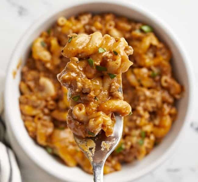 A close-up image of a spoonful of creamy macaroni and cheese with ground beef, lifted in front of a bowl filled with the dish. The macaroni is mixed with a rich, cheesy sauce and garnished with small green herbs. A striped napkin sits in the background—a perfect comfort food recipe for any occasion.