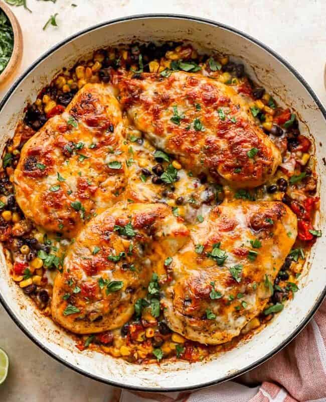 A large white skillet filled with cooked chicken breasts topped with melted cheese, surrounded by a mix of corn, black beans, and diced peppers. Fresh cilantro and lime wedges are on the side, along with a folded red and white dish towel. This dish is perfect for fans of Chicken Thigh Recipes too!