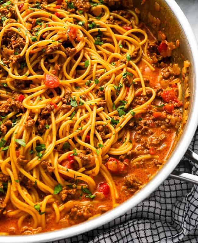 A pan filled with spaghetti cooked in a rich tomato-based meat sauce, made from cherished canned tomatoes recipes, garnished with chopped herbs. A black and white checkered cloth is draped next to the pan on the right side.