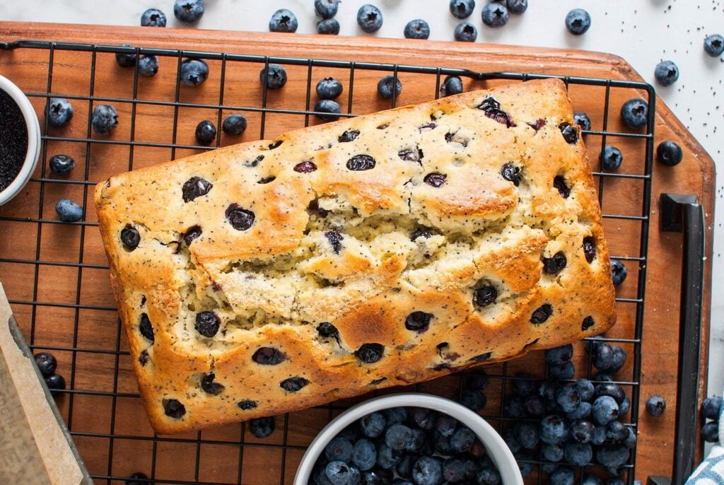 A Lemon Blueberry Poppy Seed Loaf cooling on a cooling rack next to a bowl of blueberries.