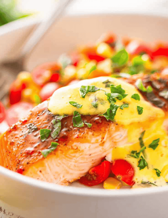 A white bowl with colorful Glazed Salmon over Corn & Tomatoes with Herb Butter Sauce.