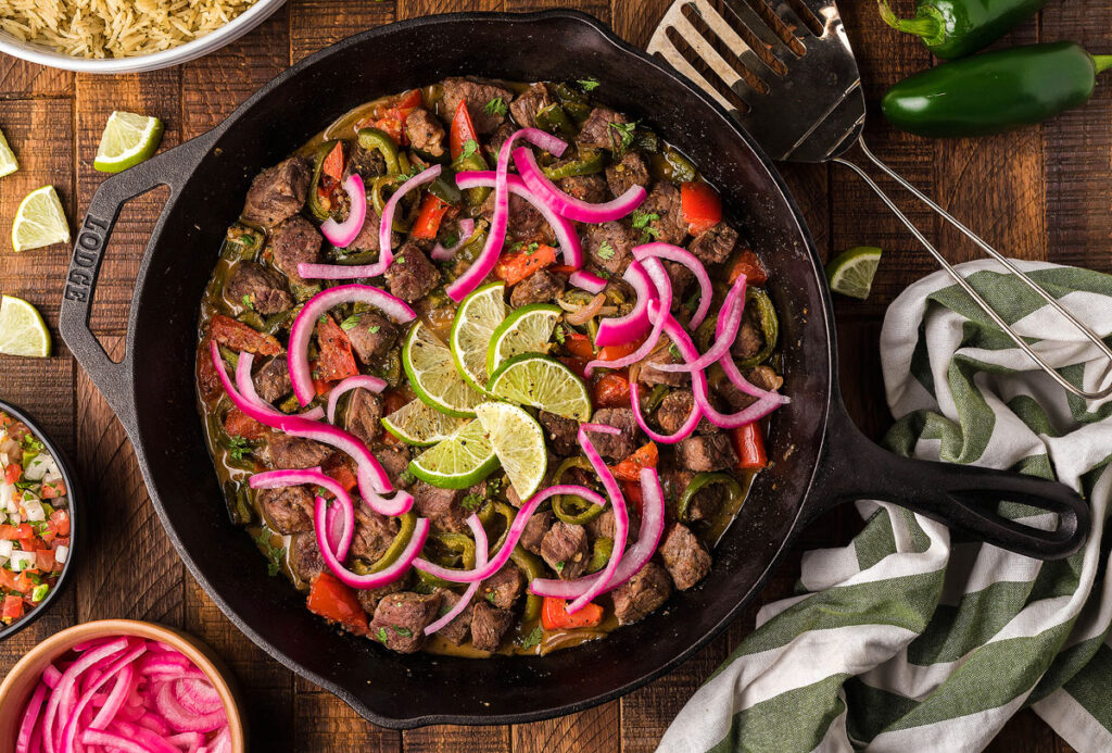 A pan of Steak Picado with red onions and lime wedges.