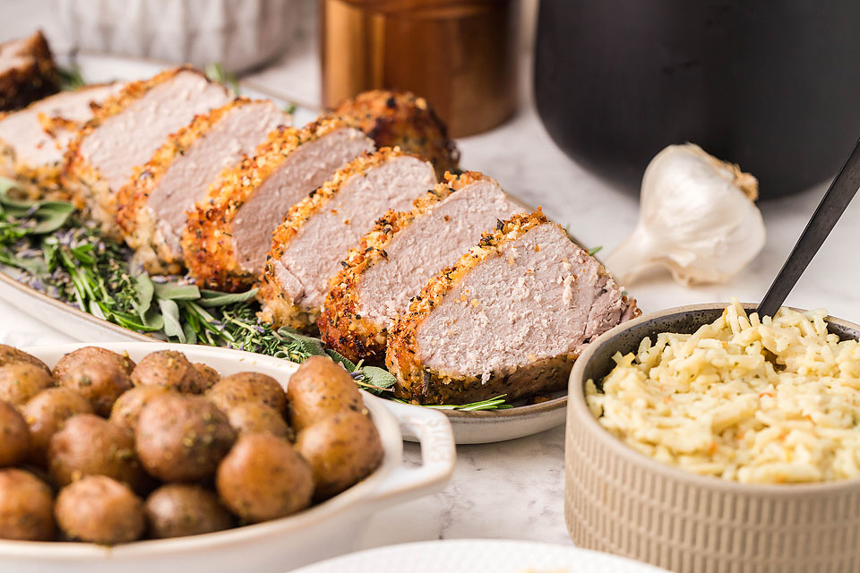 A sliced Parmesan Herb Crusted Pork Tenderloin on a platter surrounded by sides.