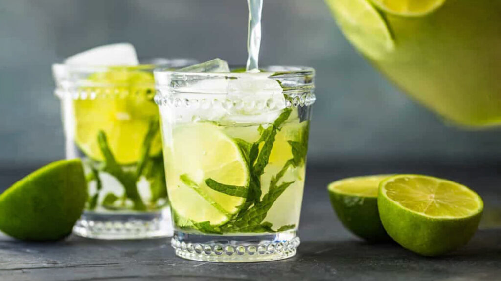 A close-up of two glasses filled with a refreshing drink, garnished with lime wedges and mint leaves. Ice cubes float in the drink as liquid is poured into one glass from a container. Lime halves are scattered around on a dark surface, perfect for exploring new cocktail recipes.