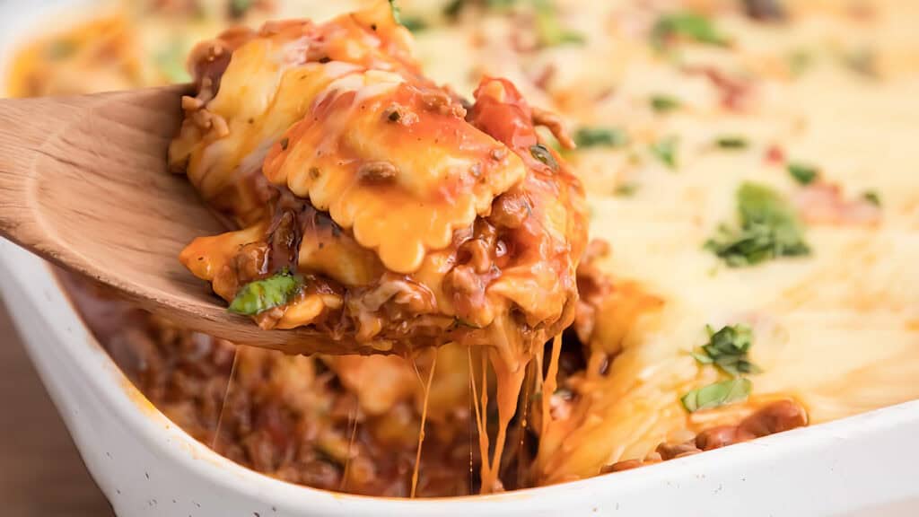 A wooden spoon lifts a heaping spoonful of cheesy Ravioli Casserole from a white baking dish. 