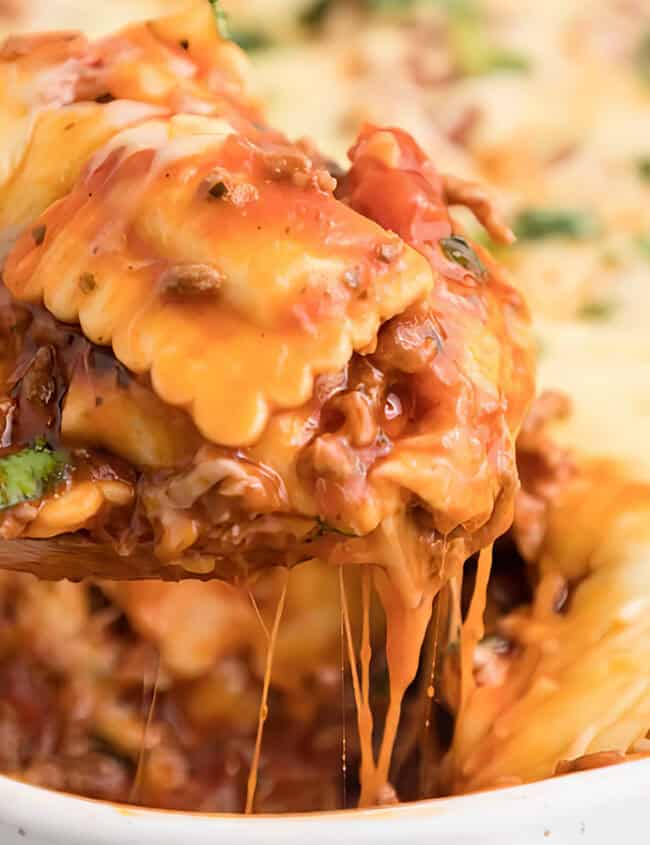 A wooden spoon lifts a heaping spoonful of cheesy Ravioli Casserole from a white baking dish.