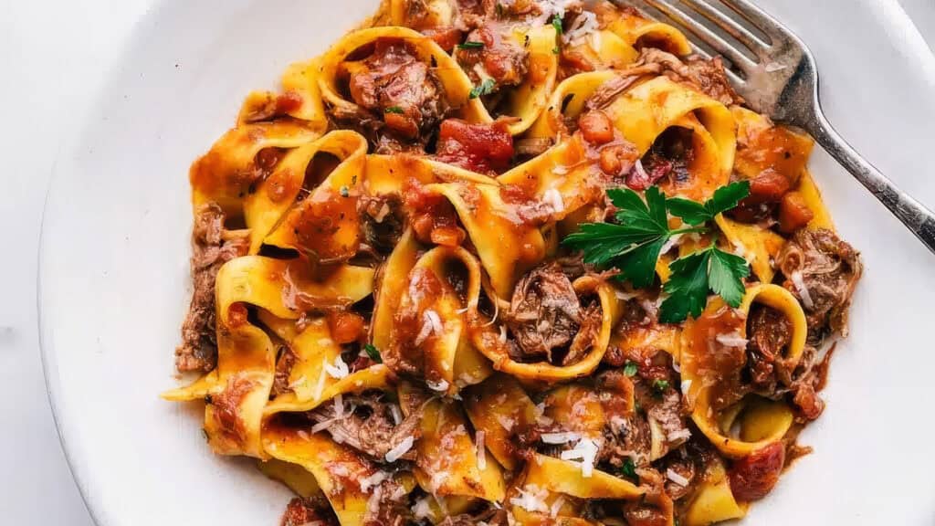 A plate of pappardelle pasta topped with a savorous beef ragu from our slow cooker recipes. The dish is garnished with a sprig of parsley and grated Parmesan cheese. A fork rests on the side of the white plate, enhancing the appetizing presentation of the rich, hearty meal.