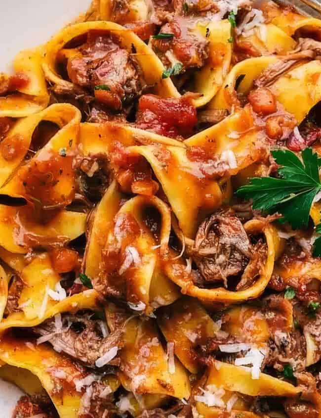 A plate of pappardelle pasta topped with a savorous beef ragu from our slow cooker recipes. The dish is garnished with a sprig of parsley and grated Parmesan cheese. A fork rests on the side of the white plate, enhancing the appetizing presentation of the rich, hearty meal.