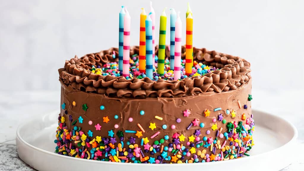 A chocolate frosted cake adorned with multicolored sprinkles and topped with six vibrant, striped candles. The sides of the cake feature a variety of star and round sprinkles, following our favorite cake recipes. The cake is presented on a white plate with a light background.