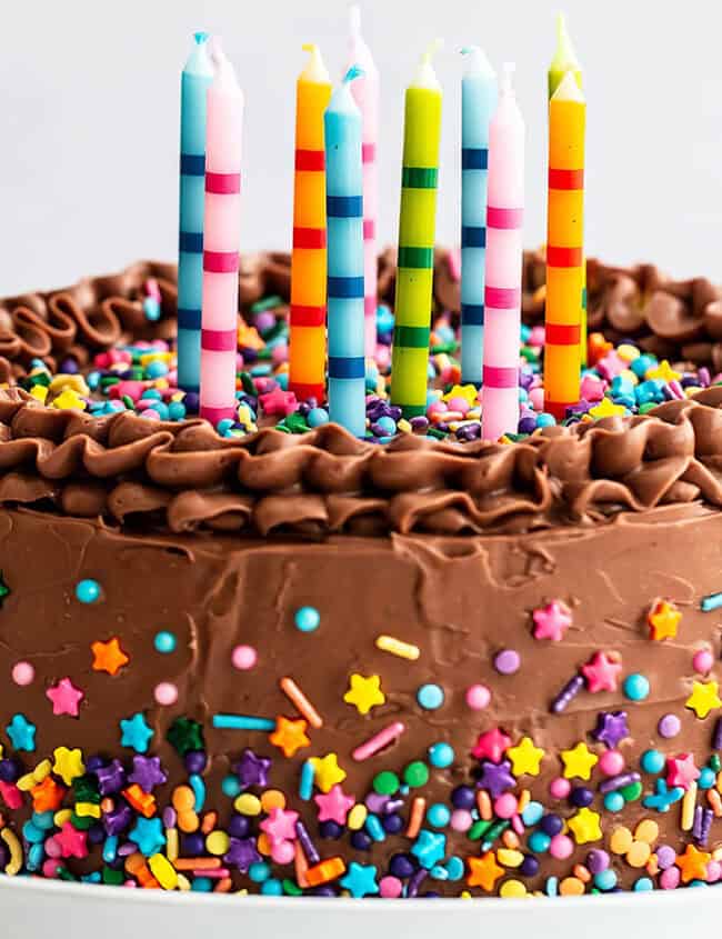 A chocolate frosted cake adorned with multicolored sprinkles and topped with six vibrant, striped candles. The sides of the cake feature a variety of star and round sprinkles, following our favorite cake recipes. The cake is presented on a white plate with a light background.