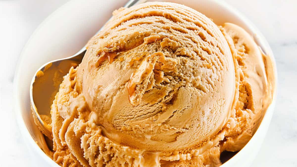 A close-up of a bowl of salted caramel ice cream with a smooth, creamy texture. A metal spoon is partially visible, scooping into the ice cream. The background is minimal, emphasizing the rich details of this delectable salted caramel ice cream recipe.