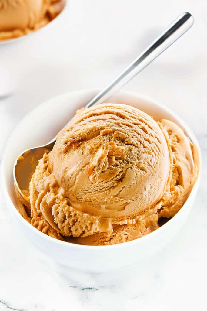 A scoop of creamy Caramel Ice Cream in a white bowl with a metal spoon beside it. This caramel dessert has a smooth, rich texture, with ridges and swirls visible. The bowl is placed on a white marble surface, creating a bright and appetizing presentation.