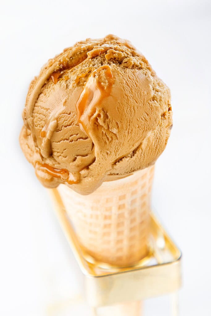 A close-up of a waffle cone filled with a single scoop of salted caramel ice cream. The ice cream is light brown with a smooth texture and has visible swirls of caramel sauce. The cone is placed upright in a holder with a blurry white background.