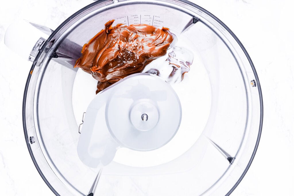 A food processor filled with creamy peanut butter and a dollop of yogurt is seen from above. The mixture, perfect for an easy recipe, appears ready to be blended, with the food processor blades visible in the center. The setup is placed on a white surface.