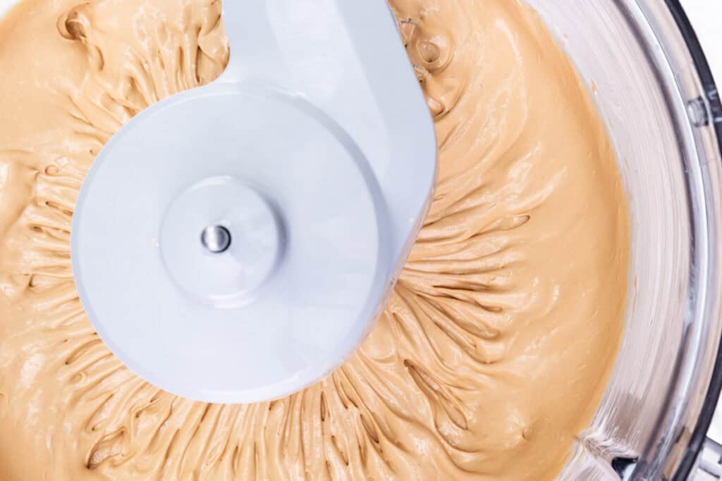 Close-up of a food processor mixing a thick, creamy, tan-colored substance, possibly dough or batter. The processor's blade is visible in the center, creating a swirling pattern in the mixture. The bowl edge is partially visible, hinting at an easy salted caramel recipe taking shape.