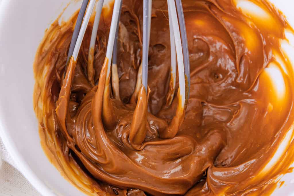 A close-up image of a metal whisk stirring thick, creamy caramel-colored mixture in a white bowl. The mixture has a smooth, glossy texture and is partially coating the whisk, perfect for your easy Salted Caramel Ice Cream recipe.