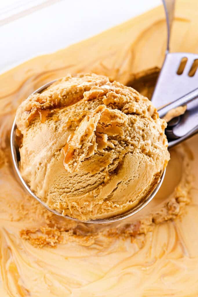 Close-up of a scoop of salted caramel ice cream in a metal scooper. The creamy ice cream has a rich caramel color with swirls of salted caramel throughout. The background shows a tub filled with more of the same salted caramel ice cream, smooth and inviting.