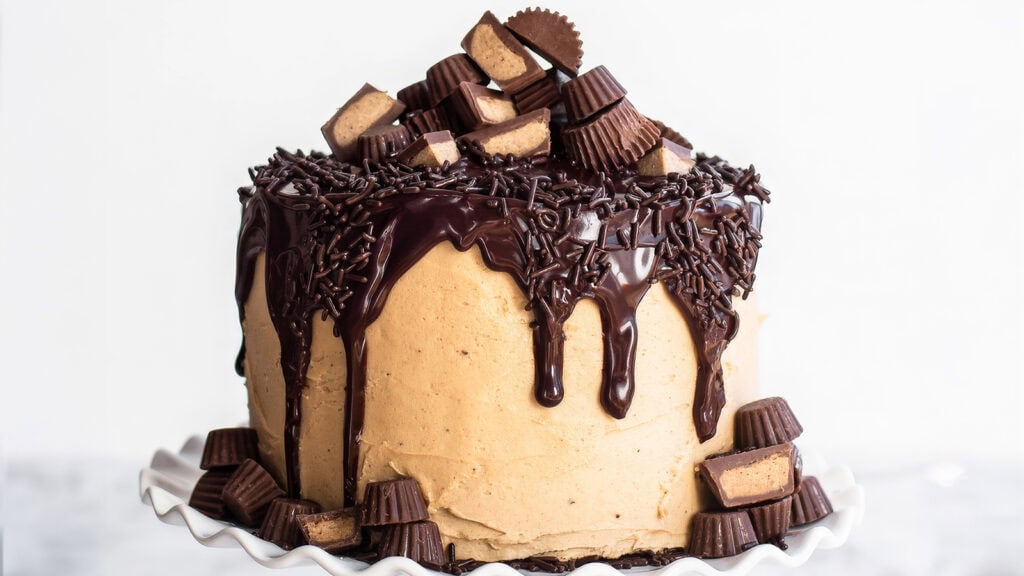 A decadent cake with smooth peanut butter frosting, topped with a generous amount of chocolate ganache dripping down the sides. The cake is adorned with chocolate sprinkles and various sizes of peanut butter cups, arranged in an appealing, stacked manner—perfect for those seeking indulgent cake recipes.