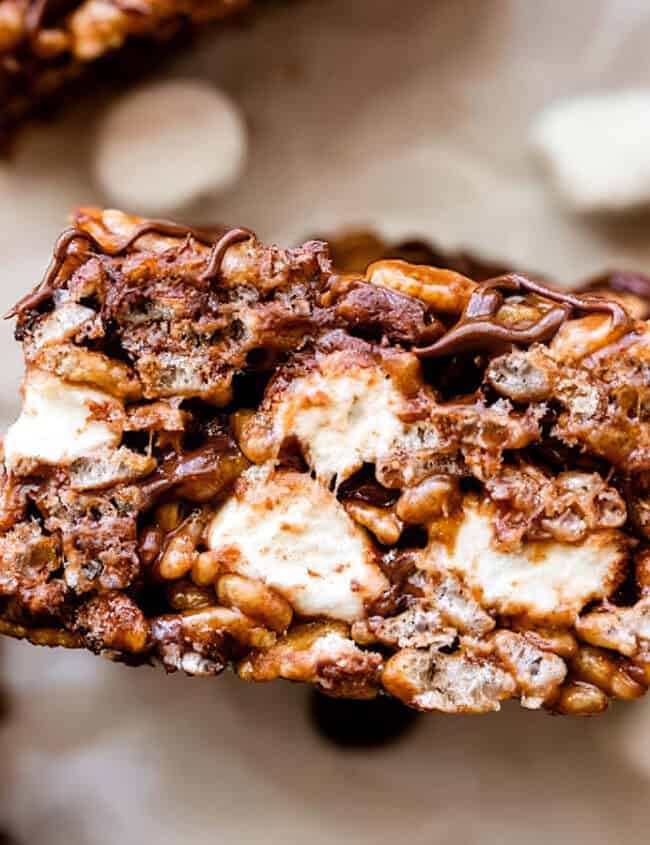 Close-up image of a chocolate rice crispy treat. The treat is filled with rice cereal and melted marshmallows, making it sticky and sweet, topped with a drizzle of chocolate. Other similar treats are partially visible in the background.