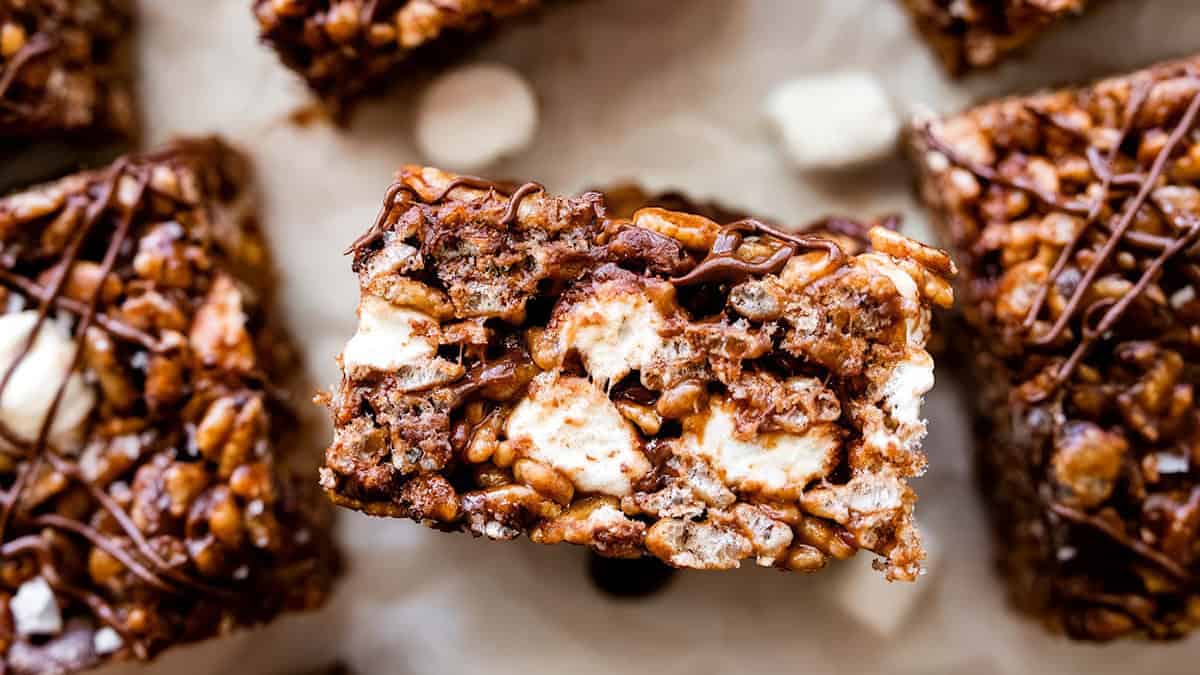 Close-up image of a chocolate rice crispy treat. The treat is filled with rice cereal and melted marshmallows, making it sticky and sweet, topped with a drizzle of chocolate. Other similar treats are partially visible in the background.