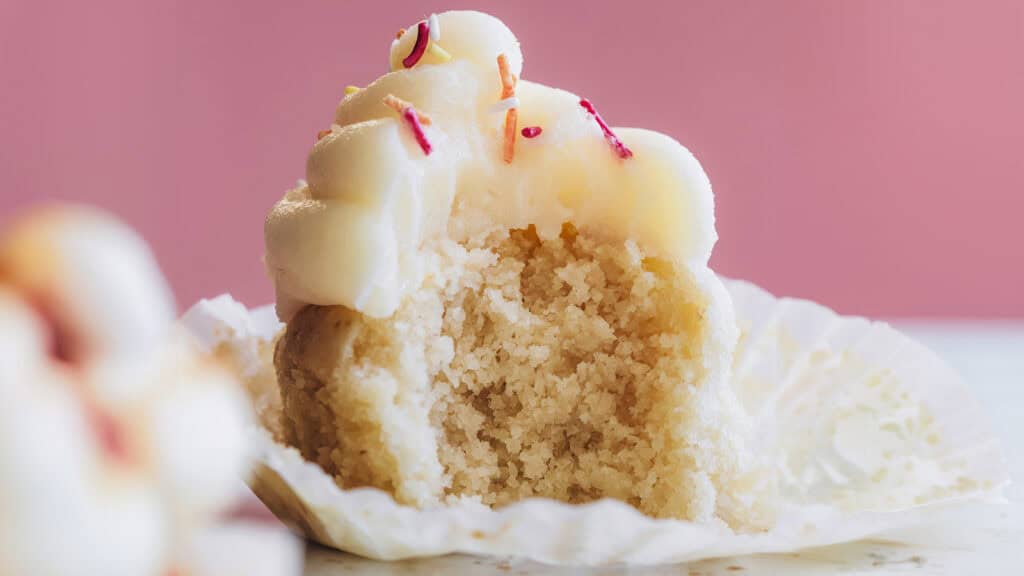 A close-up of a partially eaten vanilla cupcake with white frosting and red sprinkles. The cupcake liner is peeled back, revealing the moist, textured cake inside against a soft pink background, exemplifying the art of bakery desserts.