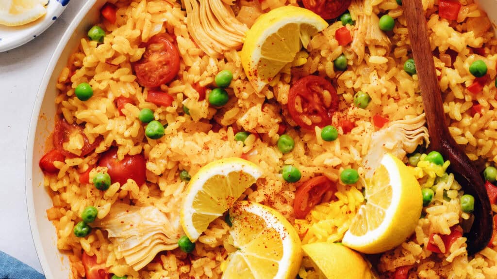 A vibrant dish of paella, featuring saffron-infused yellow rice mixed with green peas, cherry tomato slices, artichoke hearts, and garnished with lemon wedges. A wooden spoon is partially visible on the right side of the pan.