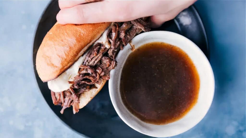 A hand holding a French dip sandwich filled with shredded beef and melted cheese above a small bowl of au jus sauce on a black plate with a light blue background, perfect for those who love Slow Cooker Recipes.