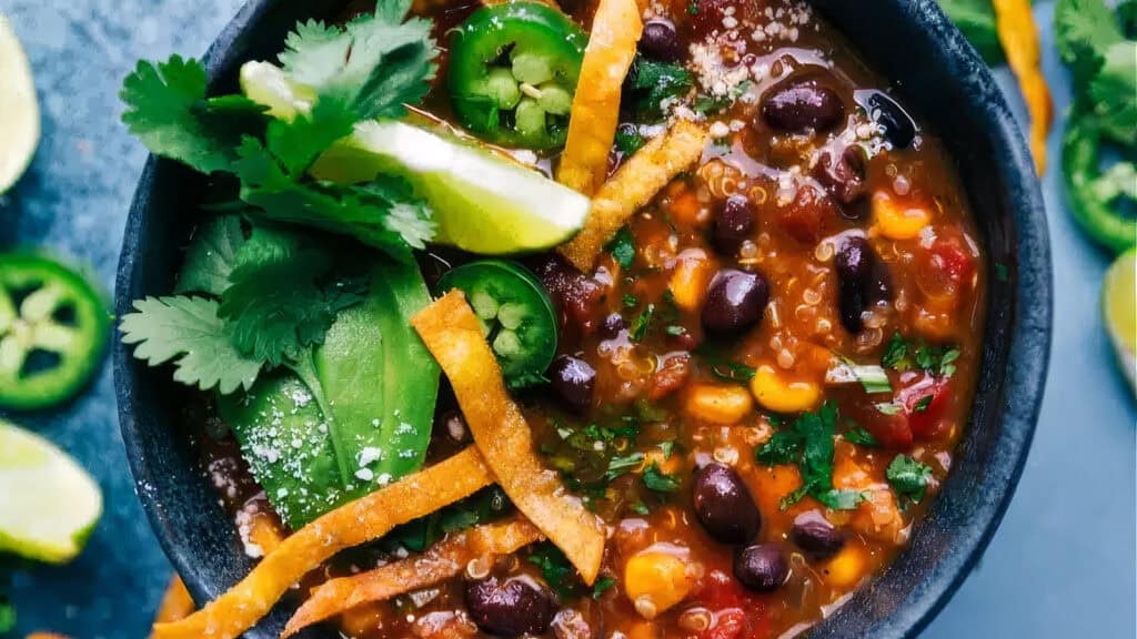 A bowl of hearty chili, perfect for any slow cooker recipe collection, is garnished with slices of avocado, lime wedges, jalapeño slices, cilantro leaves, and crunchy tortilla strips. The chili is filled with black beans, corn, tomatoes, and quinoa, creating a colorful and appetizing dish.