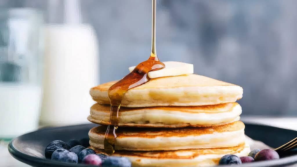 A stack of golden-brown pancakes topped with a pat of melting butter has syrup being drizzled over it. The pancakes, perfect for any pancake recipes collection, sit on a black plate surrounded by fresh blueberries. In the blurred background, a glass of milk is visible.