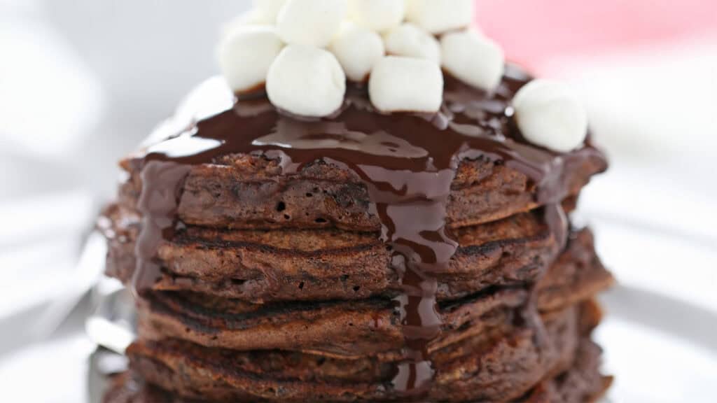 A close-up of a stack of chocolate pancakes drizzled with chocolate syrup and topped with mini marshmallows. The texture of the pancakes is visible, with a rich, dark chocolate color contrasting with the white marshmallows—a perfect highlight for your pancake recipes collection.