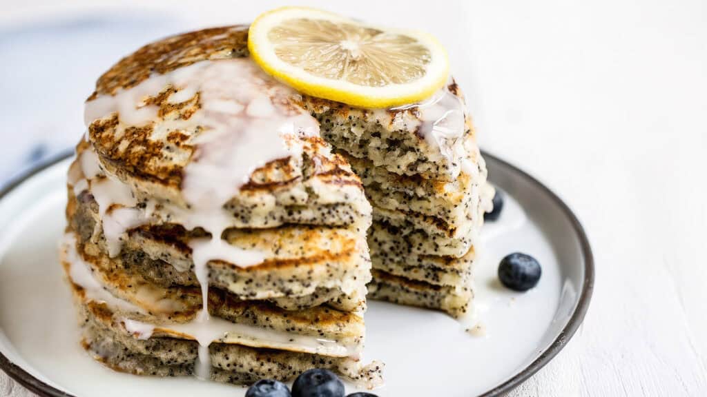 A stack of poppy seed pancakes topped with a lemon slice and drizzled with icing sits on a white plate. The pancakes have a section cut out, revealing a fluffy interior. Blueberries are scattered around the plate, presenting one of the most delightful pancake recipes against a soft white background.