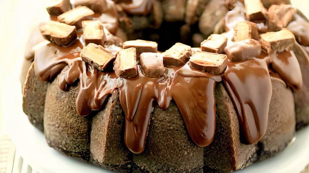 A decadent chocolate bundt cake, straight from classic bundt cake recipes, is topped with a thick layer of glossy chocolate ganache and adorned with generous chunks of chocolate bars. The rich dessert is displayed on a white plate, highlighting its indulgent and mouth-watering presentation.