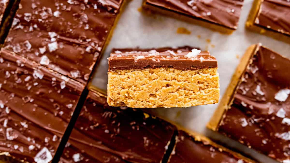 Close-up view of multiple bars of no bake peanut butter chocolate slices. The bars have a layered appearance with a thick peanut butter base and a smooth chocolate top, sprinkled with sea salt flakes. One bar is placed on its side, showcasing the thick layers.