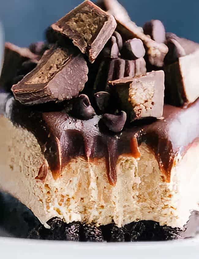 A slice of rich, no-bake peanut butter cheesecake topped with chunks of peanut butter cups and drizzled with chocolate sauce sits on a white plate. The dessert features a creamy layer over a dark cookie crust, adorned with additional chocolate pieces and sauce.