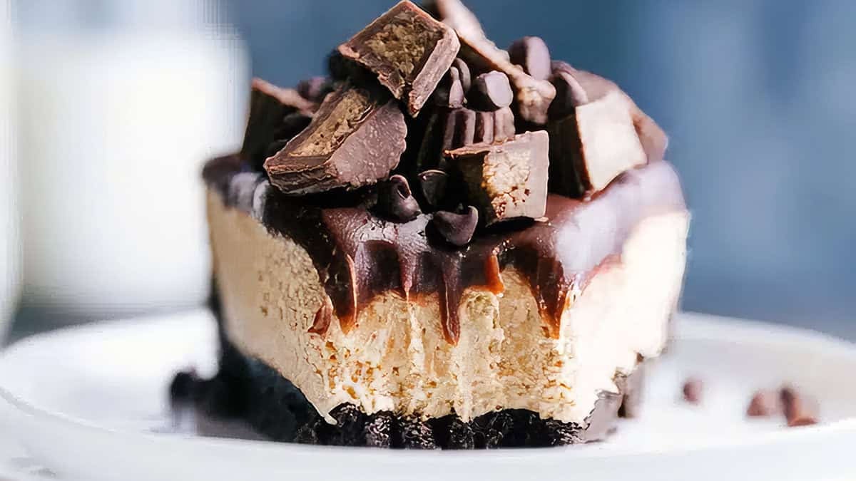 A slice of rich, no-bake peanut butter cheesecake topped with chunks of peanut butter cups and drizzled with chocolate sauce sits on a white plate. The dessert features a creamy layer over a dark cookie crust, adorned with additional chocolate pieces and sauce.