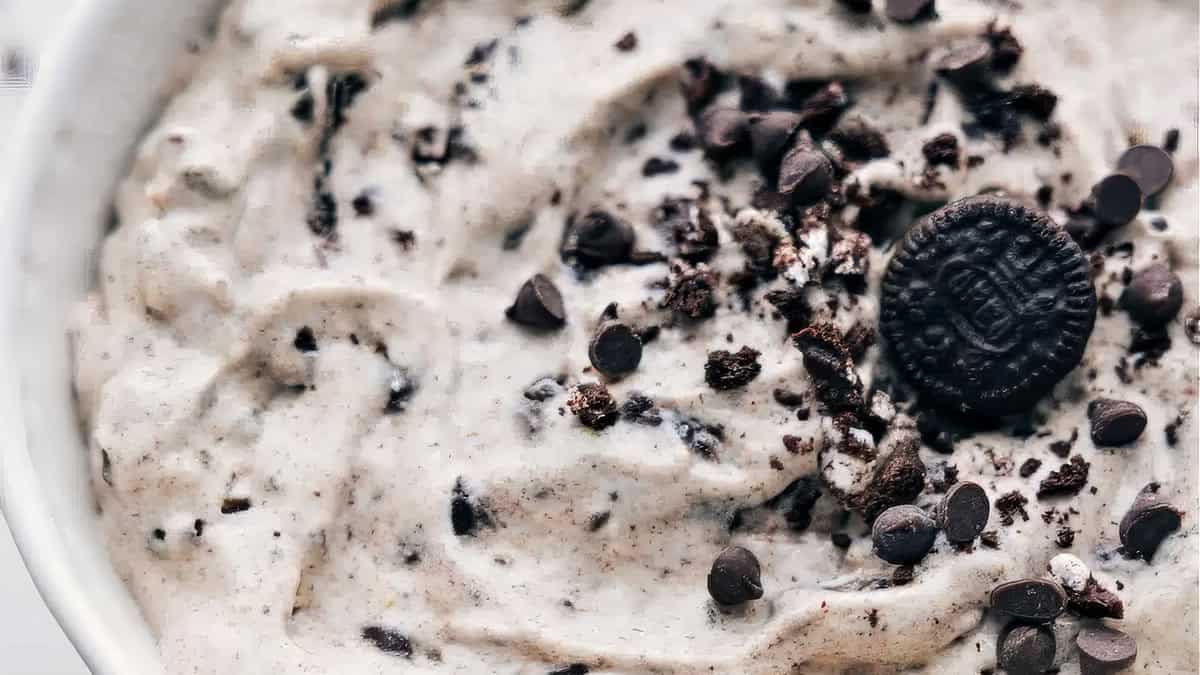 Close-up of a creamy dessert topped with crushed cookie pieces and chocolate chips. One whole chocolate sandwich cookie is visible on top, adding texture and richness to the dish. This delicious treat appears to be a cookies and cream flavored mixture.