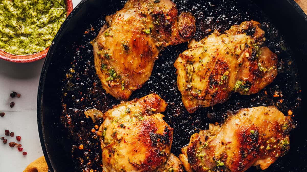 A skillet filled with four pieces of well-cooked, marinated chicken thighs, each garnished with herbs and spices. A small bowl of vibrant green pesto sauce is visible to the side, showcasing one of the best pesto recipes. The dish appears to be freshly made and flavorful.