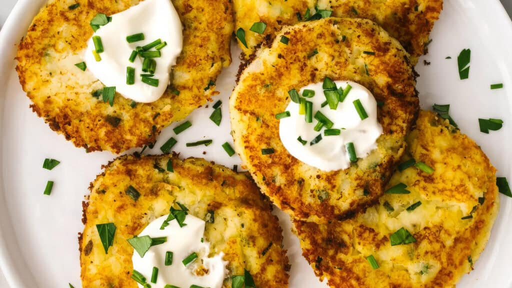 A plate of golden-brown potato pancakes topped with sour cream and garnished with fresh chopped chives. The pancakes are crispy with visible flecks of herbs, arranged neatly on a white plate—an irresistible addition to your favorite pancake recipes.